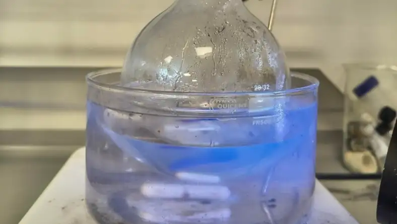 Round-bottom flask in a blue UV-lit liquid bath on a lab bench with chemistry equipment in the background