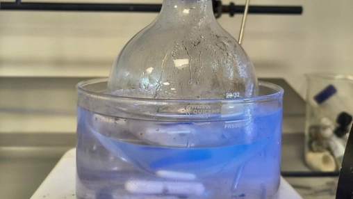 Glass round-bottom flask submerged in a blue ice bath inside a clear plastic container on a lab bench