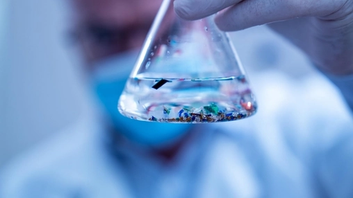 Close-up of a glass conical flask held by a gloved hand, containing colorful plastic particles in clear liquid