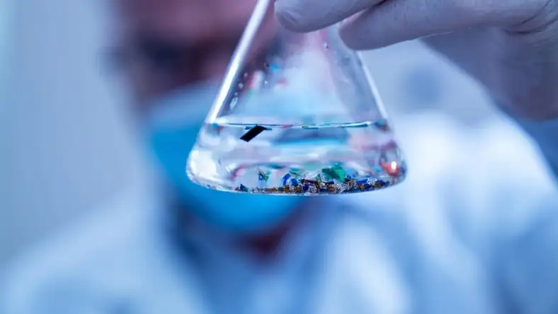 Close-up of a glass conical flask held by a gloved hand, containing colorful plastic particles in clear liquid