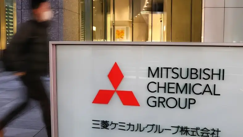 Mitsubishi Chemical Group entrance sign with Japanese text, a pedestrian blurred in motion passing behind