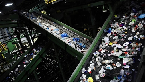 Conveyor belts crisscross a recycling facility, carrying mixed plastic waste past green metal support frames