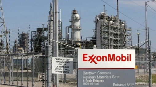 An ExxonMobil sign at the Baytown Complex refinery entrance with industrial towers and pipework behind a fence