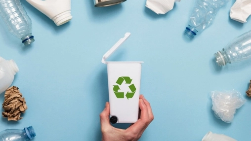 A hand holds a small white bin with a recycling symbol surrounded by scattered plastic bottles and paper