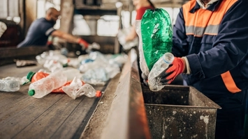 Two workers in high-visibility jackets sorting plastic bottles on a conveyor belt at a recycling facility