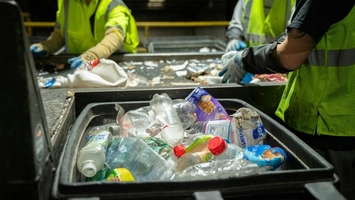 Workers in yellow safety vests and blue gloves sorting plastic bottles and packaging at a recycling facility