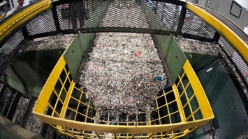 Mixed plastic waste on a conveyor belt with yellow safety barriers at the sides inside a recycling facility