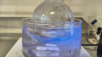 Round-bottom flask in a blue UV-lit liquid bath on a lab bench with chemistry equipment in the background